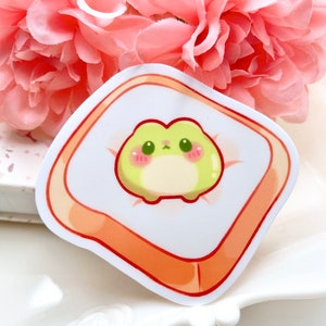 May include: A sticker featuring a cartoon frog with big eyes and a pink blush, sitting on a slice of toast. The frog is green and yellow, and the toast is orange.