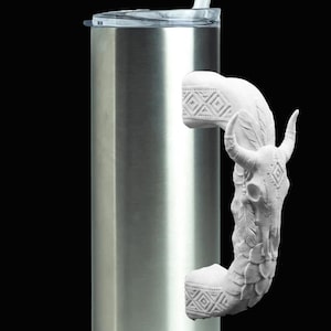 May include: A tall, cylindrical stainless steel tumbler with a clear plastic lid and a white straw. The handle is a white, sculpted 3D figure of a bull skull adorned with feathers and geometric patterns, attached to the side of the metal cup.