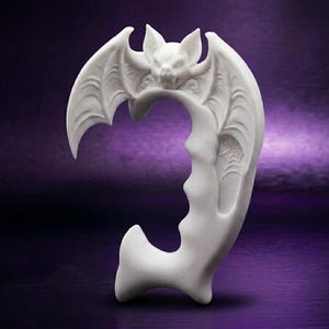 May include: A white, C-shaped ear cuff featuring a sculpted bat at the top with spread wings. The design includes intricate details on the wings and a small spiderweb pattern on the lower section, creating a curved, solid structure for the ear.