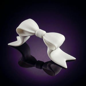 May include: A solid white decorative bow ornament featuring smooth, curved loops and two pointed, flowing ribbon ends. The object has a matte finish and is shaped to resemble a tied fabric bow, with a central knot connecting the two symmetrical loops.