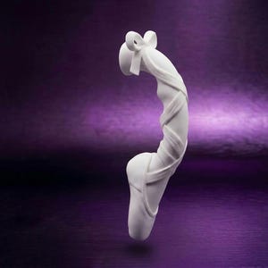 Puede incluir: Una escultura de color blanco mate con forma de zapatilla de ballet con cintas que se envuelven alrededor del área del tobillo. La zapatilla presenta una puntera redondeada y un pequeño detalle de lazo en la parte superior de las cintas.