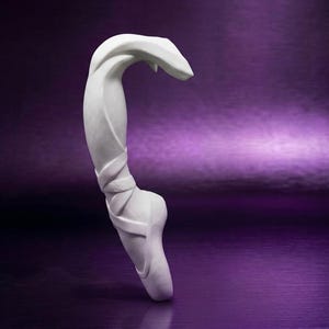 Puede incluir: Una escultura blanca de una zapatilla de ballet con cintas envueltas alrededor del área del tobillo. La zapatilla está colocada verticalmente, mostrando la puntera estructurada y las líneas curvas y fluidas del material con acabado mate.