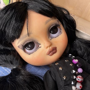 May include: A black-haired doll with large, white eyes and dark eye makeup. The doll is wearing a black jacket with silver stars and a beaded necklace with purple, white, and black beads.