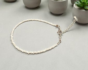 May include: A delicate white beaded bracelet with a silver clasp and chain. The bracelet is made of small, round, white beads. The clasp and chain are silver-coloured. The bracelet is shown on a grey surface.