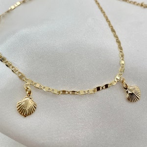 Könnte beinhalten: Ein goldenes Fußkettchen mit zwei kleinen Muschel-Anhängern. Das Fußkettchen hat einen Karabinerverschluss und eine verstellbare Schiebekugel.