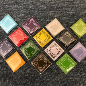 May include: Twelve decorative ceramic tiles with a geometric pattern in various colors, including blue, red, green, yellow, purple, and pink. The tiles are arranged in a diamond shape on a dark blue denim background.