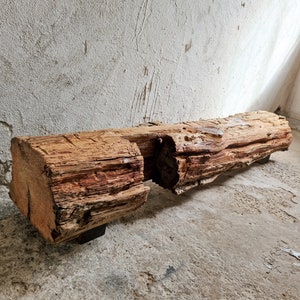 Puede incluir: Un banco de madera rústico hecho de un tronco grande y desgastado con nudos y vetas naturales. El banco tiene una superficie rugosa y desigual y está sostenido por dos patas de metal negro.