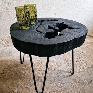 May include: A black wooden coffee table with a unique, natural wood grain pattern and a live edge design. The table has three hairpin legs and is sitting on a concrete floor.