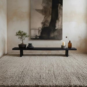 May include: A minimalist interior scene featuring a black bench with a bonsai tree, stacked stones, candles, and a small vase. A large abstract painting hangs above the bench. A textured, neutral-toned rug covers the floor.