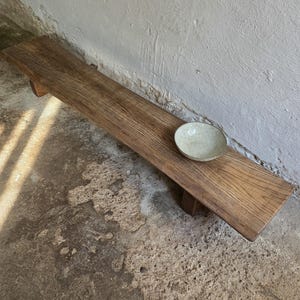 May include: A long, low-profile wooden bench with a natural grain finish, supported by short block legs. A single, shallow, off-white ceramic bowl with a speckled texture rests on the right side of the bench surface, showcasing a simple design.