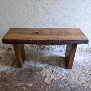 May include: A rectangular wooden bench featuring a thick, dark brown slab seat with a visible natural wood grain and a prominent knot. The bench is supported by two sturdy, vertical wooden legs that extend to the floor, providing a stable base.