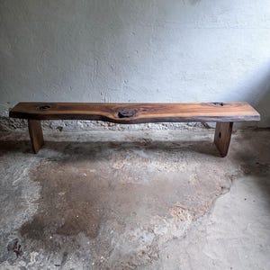 Könnte beinhalten: Eine lange, rechteckige Holzbank mit natürlicher Baumkante und einer satten braunen Holzmaserung. Die Sitzfläche weist drei markante dunkle Astlöcher auf und wird von zwei stabilen, flachen Holzbeinen getragen, von denen eines ein Loch hat.