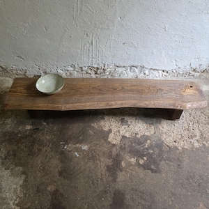 Op de afbeelding: Een lage, rechthoekige houten bank met een natuurlijke boomkant en zichtbare houtnerf, ondersteund door korte blokpoten. Een kleine, ronde, lichtgekleurde keramische kom met een gespikkelde afwerking staat aan de linkerkant van de bank.
