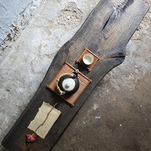 Op de afbeelding: Een donker gebeitste houten plank met natuurlijke rand, voorzien van een zwarte keramische theepot met bamboe handvat, een klein wit keramisch kopje op een bamboe matje, en een stuk verouderd papier met een gedroogde bloemstengel.