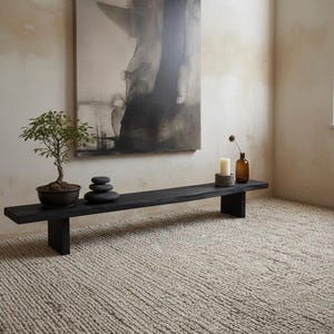 Pode incluir: Um banco comprido de madeira preta com borda natural, contendo um pequeno bonsai verde num vaso escuro, uma pilha de três pedras pretas lisas, uma vela branca sobre uma base de pedra cinzenta e uma pequena garrafa de vidro castanho com uma flor.