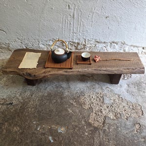 Op de afbeelding: Een lage, rechthoekige houten tafel met een natuurlijke boomkant, voorzien van een donkere keramische theepot met bamboe handvat op een bamboemat, een klein keramisch kopje op een vierkante onderzetter, een stuk gestructureerd papier en een gedroogde bloemstengel.