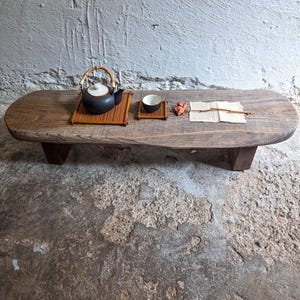 Puede incluir: Una mesa baja rectangular de madera con bordes redondeados y cuatro patas en bloque. Sobre la superficie hay una tetera de cerámica negra con asa de bambú sobre una esterilla, una pequeña taza de té a juego y una flor seca sobre un papel.