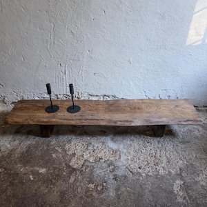 Op de afbeelding: Een lage, rechthoekige houten bank met een natuurlijke boomkant en een donkerbruine houtnerfafwerking. Twee slanke, matzwarte metalen kandelaars met cilindrische toppen staan aan de linkerkant van het oppervlak van de bank.