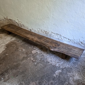 May include: A long, low-profile wooden bench featuring a dark brown stained finish and a natural live edge along the front. The bench is supported by two short, rectangular wooden legs and displays a prominent wood grain pattern throughout the surface.
