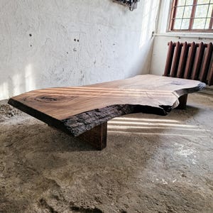 May include: A large, natural-edge wooden coffee table. The tabletop is a single slab of light brown wood with visible grain and a live edge. It rests on a dark brown, geometric base. The table is in a room with a concrete floor and a window.