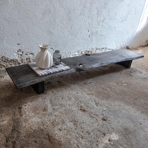 May include: A low, rectangular wooden bench with a dark, charred finish and a natural live edge. A white, ribbed ceramic vase and a small, clear glass bottle sit on top of a book placed on the left side of the bench surface.