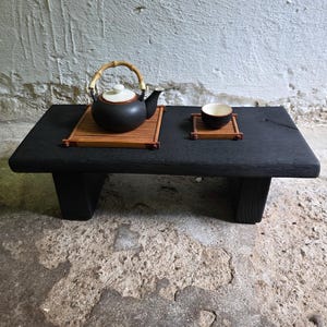 Op de afbeelding: Een lage zwarte houten tafel met een matte afwerking, voorzien van een donkere keramische theepot met een bamboe handvat en een bijpassend keramisch theekopje. Beide items staan op individuele rechthoekige bamboematten met houten frames.