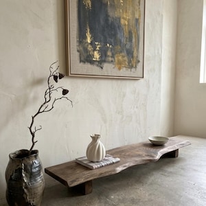 May include: A rustic, live-edge wooden bench with a natural, irregular shape. A white, textured vase sits on a book, and a small bowl is placed on the bench. A large, abstract painting with gold and blue tones hangs on the wall.