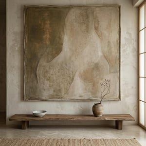 May include: A large square framed abstract painting with muted beige, tan, and olive green tones hangs above a long, low wooden bench. On the bench sits a small ceramic bowl and a rounded clay vase containing a single thin, dried branch.