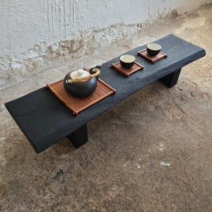 Op de afbeelding: Een zwarte houten theetafel met een laag profiel, met een theeset. De set bevat een donkerbruine theepot met een wit deksel, twee kleine theekopjes en bamboe serveerbladen. De tafel is rechthoekig en heeft twee poten.