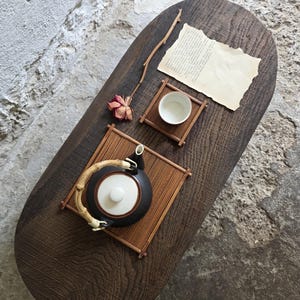 Op de afbeelding: Een donkerbruine ovale houten tafel met een zwarte theepot met wit deksel en bamboe handvat, een klein wit theekopje, twee bamboe matjes, een gedroogde roos en een stuk verouderd papier met gedrukte tekst op het oppervlak.