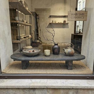 Può includere: Vetrina di "Wabi Sabi Ceramics". Un tavolo in legno scuro espone una ciotola in ceramica, una tazza, un vaso e una teiera. Un ramo secco è nel vaso. Scaffali sullo sfondo mostrano ceramiche e tessuti.