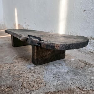 May include: A dark, rustic wooden coffee table with a live edge design. The table has a rectangular top with a natural, uneven edge and sits on two solid, block-shaped legs. The wood has a dark, weathered appearance.