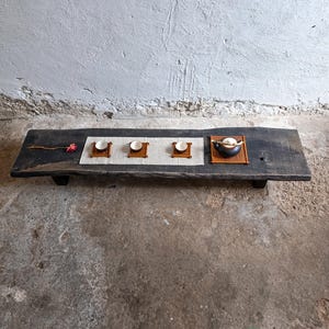 May include: A rustic wooden tea table with a dark finish. It features three small white tea cups on individual wooden stands, a small black teapot, and a decorative flower. The table is set on a concrete floor.