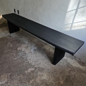 May include: A long, black wooden bench with a flat, rectangular seat and two sturdy, vertical slab legs. Two thin, black metal candle holders are positioned on the left side of the bench surface, which displays a visible wood grain texture.