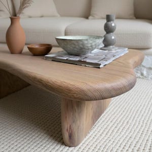 Puede incluir: Una mesa de centro de madera marrón claro con un diseño curvo único. La mesa presenta una veta de madera natural y se apoya sobre una alfombra color crema. Los elementos decorativos incluyen un jarrón de cerámica, un cuenco de madera y un jarrón gris.