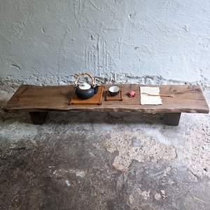 Puede incluir: Una mesa baja rectangular de madera con borde natural y acabado mate oscuro. Sobre la superficie hay una tetera de cerámica negra con asa de bambú sobre una bandeja de bambú, una pequeña taza de té a juego, una flor seca y un trozo de papel.