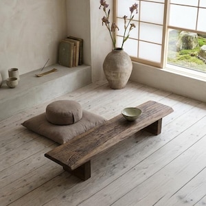 May include: A low rectangular wooden bench with a weathered texture, featuring a small ceramic bowl on its surface. Beside the bench sits a square floor cushion topped with a round meditation pillow, all set on light-coloured wooden floorboards.