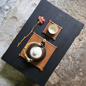 Könnte beinhalten: Eine schwarze Keramik-Teekanne mit weißem Deckel und gebogenem Bambusgriff, eine kleine weiße Keramiktasse und zwei rechteckige Bambusmatten. Eine einzelne getrocknete rosa Rose an einem dünnen Stiel liegt neben der Teekanne auf Holz.