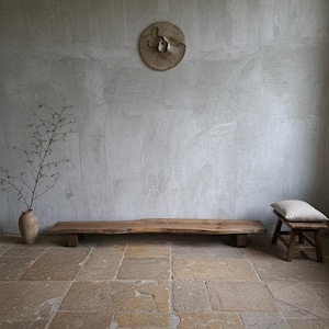 May include: A long, natural-edged wooden bench on a stone floor. A small wooden stool with a cushion is to the right. A vase with a plant and a circular wall hanging are also visible, creating a minimalist aesthetic.