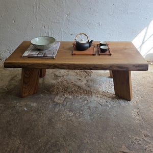 Peut inclure: Une table basse rectangulaire en bois avec un bord naturel, présentant un bol en céramique claire sur un magazine, une théière sombre avec une anse en bambou sur un plateau en bois, et deux petites tasses assorties sur un plateau séparé.