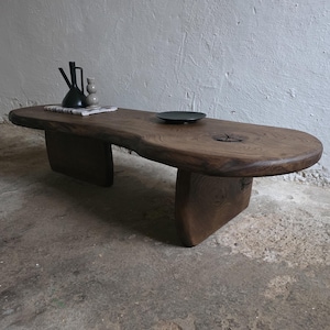 Pode incluir: Uma grande mesa de centro de madeira maciça com uma forma natural e orgânica. A mesa castanha escura apresenta uma borda viva e duas pernas retangulares grossas. Um prato preto e vasos decorativos estão em cima. A mesa tem aproximadamente 183 cm de comprimento.