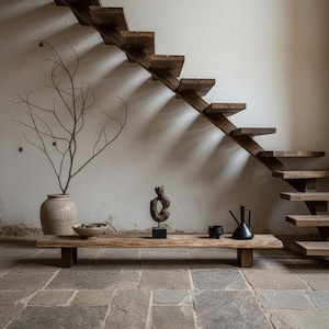 Op de afbeelding: Een rustiek interieur met een houten trap en een lage, rechthoekige houten tafel. De tafel toont decoratieve items, waaronder een sculptuur, een kom en een zwarte kan. Er staat ook een grote vaas met takken.