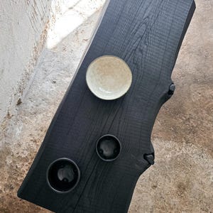 May include: A black, live-edge wooden table with a natural, organic shape. The table features three circular indentations and a small, speckled bowl. The wood grain is visible, and the table has a matte finish.
