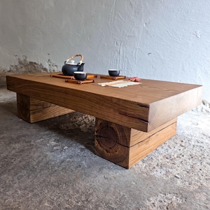 Op de afbeelding: Een lage rechthoekige salontafel gemaakt van dik, massief bruin hout met een zichtbare nerf. De tafel heeft twee stevige blokvormige poten. Op het oppervlak staan een zwarte theepot met een bamboe handvat, twee kleine zwarte kopjes en een stukje papier.