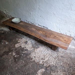 Könnte beinhalten: Eine lange, niedrige Holzbank mit natürlicher Baumkante und einer reichen braunen Holzmaserung. Die Bank wird von zwei kurzen, blockartigen Beinen gestützt und trägt eine einzelne, kleine, helle Keramikschale auf ihrer Oberfläche.