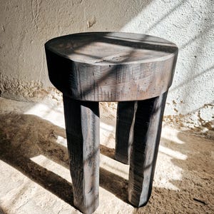 Puede incluir: Un taburete de madera en color gris carbón oscuro con un asiento circular grueso y tres patas cilíndricas robustas. La madera muestra una textura de grano visible y tosca con un acabado mate, exhibiendo imperfecciones naturales y un aspecto desgastado.