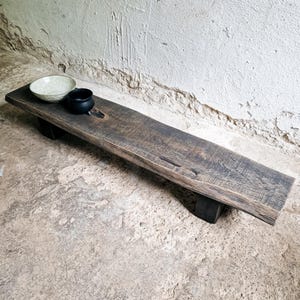 Può includere: Un lungo vassoio da portata in legno scuro con un'estetica naturale e rustica. Il vassoio rettangolare è sostenuto da due blocchi neri. Due piccole ciotole in ceramica, una chiara e una nera, sono posizionate sul vassoio.