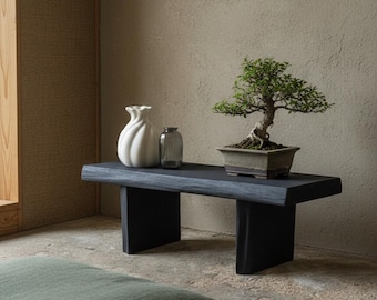 May include: A dark gray wooden bench with a bonsai tree in a square pot, a white vase, and a small glass bottle. The bench is set against a neutral-toned wall. A green cushion is on the floor.