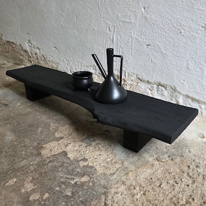 May include: A black wooden serving tray with a live edge, holding a small black bowl and a black pitcher with a handle. The tray is supported by two rectangular legs. The objects are displayed against a textured, light gray wall.