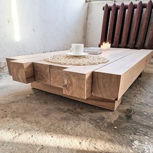 Puede incluir: Mesa de centro de madera clara con diseño geométrico. La mesa tiene una parte superior rectangular y patas gruesas de corte cuadrado. Una taza y un platillo blancos se colocan sobre un salvamanteles tejido. También hay velas y un libro sobre la mesa.